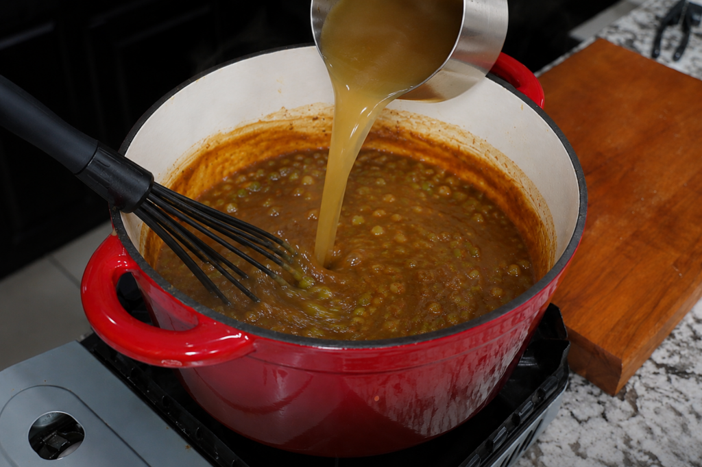 A red pot of lentil soup cooks on a stove as broth is poured in from a metal measuring cup; a black whisk rests inside the pot, and a wooden cutting board sits nearby.