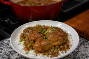 A white plate of rice topped with two seasoned chicken drumsticks, peas, and vegetables in sauce. In the background, there is a red pot with more chicken on a stovetop.