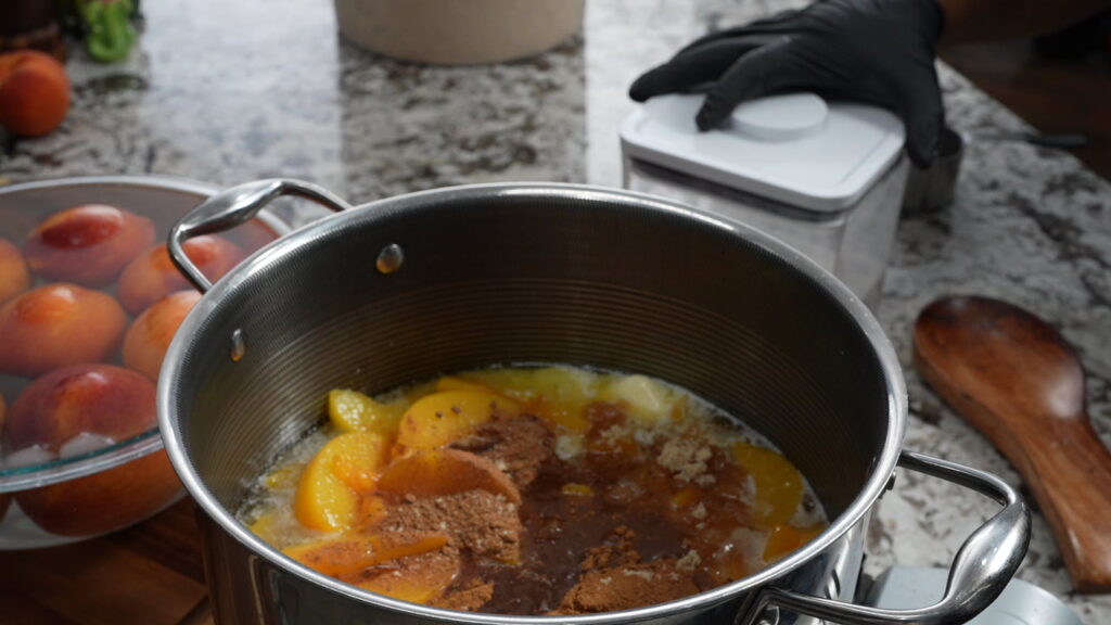 A pot with peaches, spices, and liquid sits on a counter, ready to become the base for a delicious peach cobbler. A gloved hand holds a white container nearby. Whole peaches and a wooden spoon are also visible on the granite countertop.