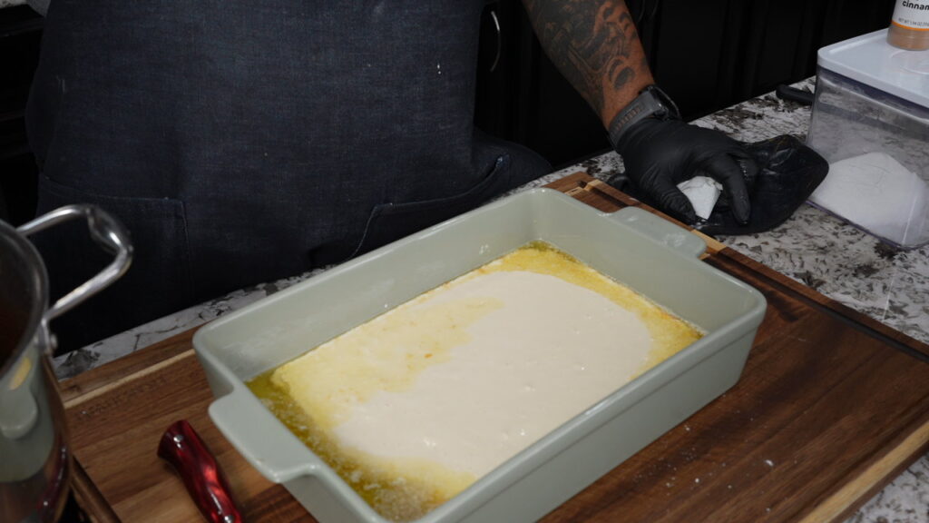 A person wearing a dark apron and black gloves is preparing food, carefully pouring batter for peach cobbler into a rectangular baking dish on a wooden surface. Nearby, kitchen utensils and containers are visible.