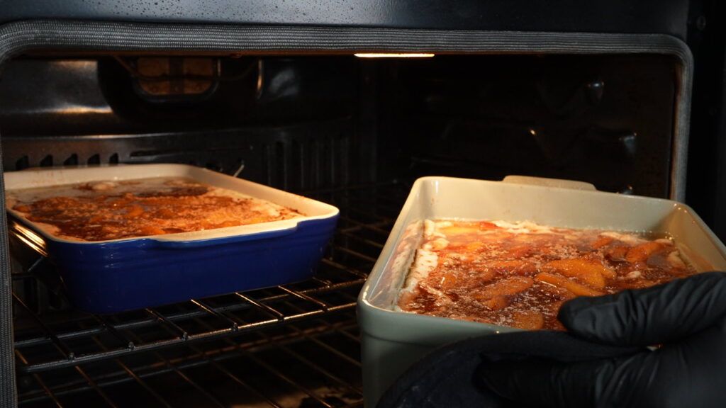 Two casserole dishes filled with peach cobbler are baking in an oven. A person wearing a black oven mitt is holding the edge of one dish.