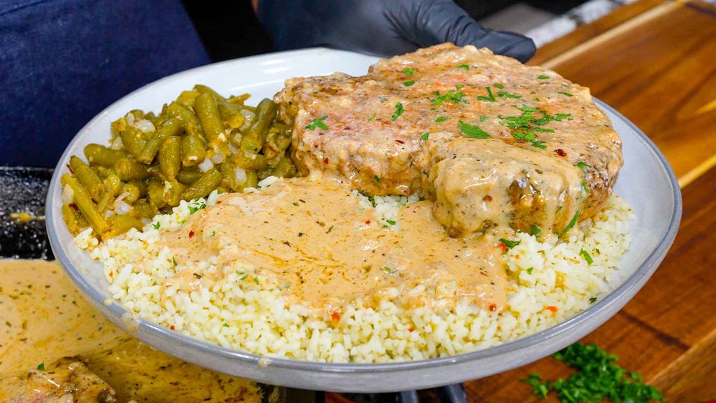 A plate with creamy garlic parmesan pork chops smothered in rich gravy, white rice topped with savory sauce, and seasoned green beans, held by a gloved hand above a wooden surface.