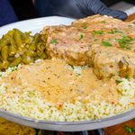 A plate with creamy garlic parmesan pork chops smothered in rich gravy, white rice topped with savory sauce, and seasoned green beans, held by a gloved hand above a wooden surface.