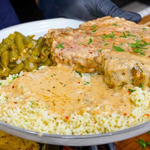 A plate with creamy garlic parmesan pork chops smothered in rich gravy, white rice topped with savory sauce, and seasoned green beans, held by a gloved hand above a wooden surface.