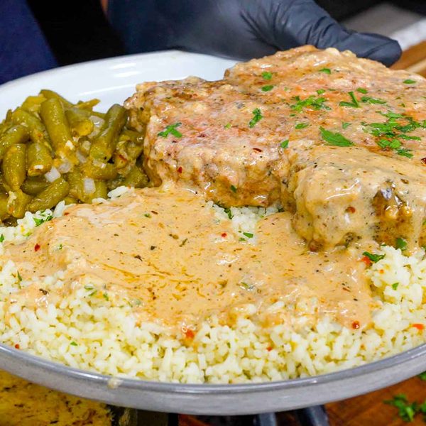 A plate with creamy garlic parmesan pork chops smothered in rich gravy, white rice topped with savory sauce, and seasoned green beans, held by a gloved hand above a wooden surface.