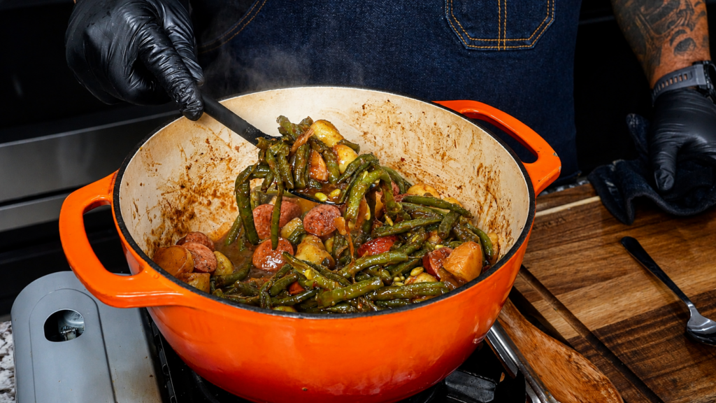 A person wearing black gloves stirs smothered green beans with potatoes and sausage in a large orange Dutch oven on a stove, with steam rising from the food.