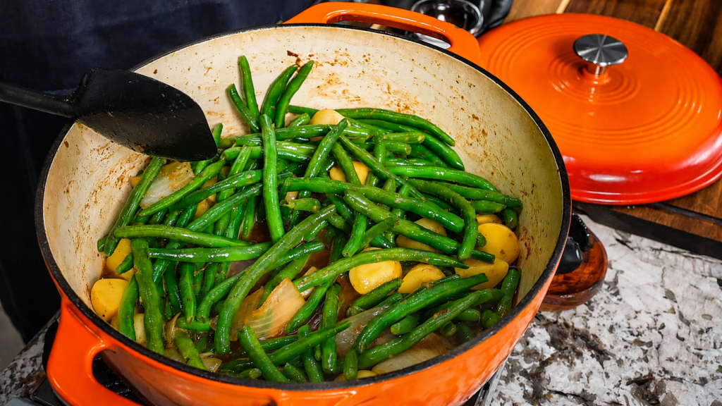 A pot of smothered green beans with potatoes and sausage, along with onions, is being cooked, with a black spatula resting on the side. The pot is orange with a lid set beside it on a granite countertop.