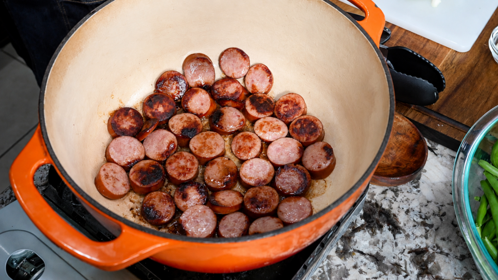 Sliced sausage pieces are being browned in a large orange Dutch oven on a stovetop, with a bowl of green beans and other ingredients nearby—perfect for preparing smothered green beans with potatoes and sausage.