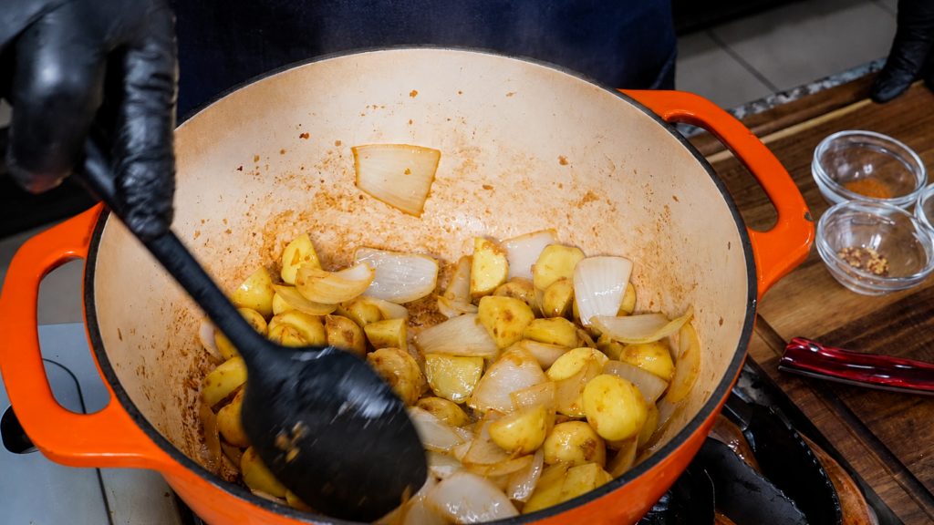 A person wearing black gloves stirs diced potatoes and onions in an orange pot on a stove, preparing the base for smothered green beans with potatoes and sausage. Two small glass bowls sit on a wooden countertop nearby.