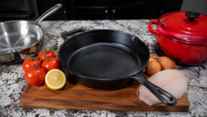 A black cast iron skillet on a wooden board with tomatoes, half a lemon, eggs, and raw fish nearby, all prime examples of what not to cook in cast iron if you want to preserve your cookware. A red Dutch oven and stainless steel pan sit on a granite countertop in the background.