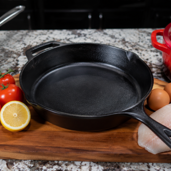 A black cast iron skillet on a wooden board with tomatoes, half a lemon, eggs, and raw fish nearby, all prime examples of what not to cook in cast iron if you want to preserve your cookware. A red Dutch oven and stainless steel pan sit on a granite countertop in the background.
