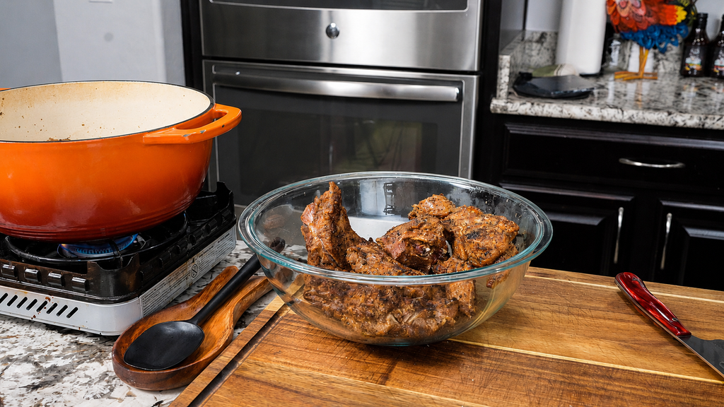 A glass bowl with cooked, seasoned smothered neck bones sits on a wooden cutting board next to a red Dutch oven on a stove in a modern kitchen. A wooden spoon and red-handled knife are also on the counter.