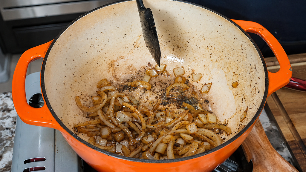 Chopped onions being sautéed in a large orange Dutch oven on a stovetop, with a black spatula resting inside. Light browning and seasoning are visible on the onions and pot, forming the aromatic base for classic smothered neck bones.