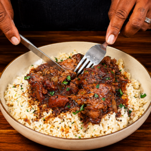 A person uses a fork and knife to cut into braised smothered neck bones served over rice on a plate, garnished with herbs, on a wooden table.