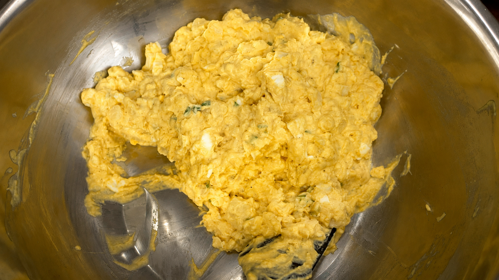 A bowl of yellow, chunky egg salad mixture with bits of herbs and a hint of potato salad in a large stainless steel mixing bowl.