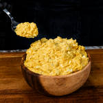 A person wearing black gloves holds a spoonful of creamy potato salad above a wooden bowl brimming with the classic yellow potato salad, all set atop a rustic wooden cutting board.