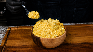 A person wearing black gloves holds a spoonful of creamy potato salad above a wooden bowl brimming with the classic yellow potato salad, all set atop a rustic wooden cutting board.