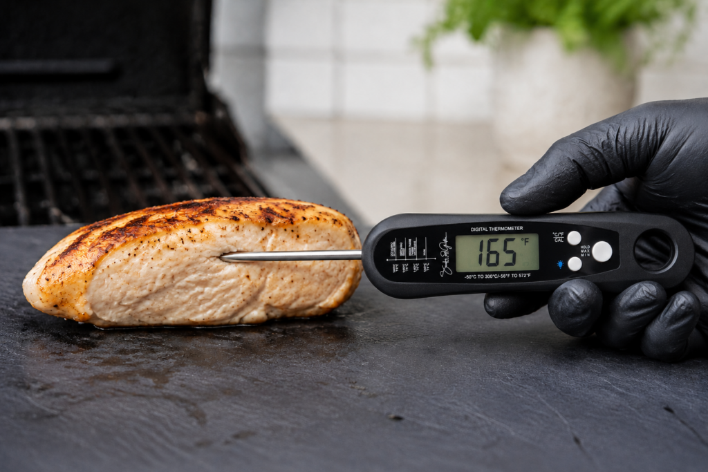 A gloved hand demonstrates how to use a meat thermometer by checking a cooked chicken breast, displaying a temperature of 165.7°F, beside a grill.