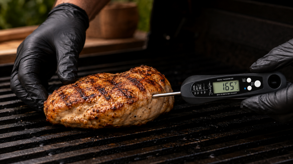 A gloved hand holds grilled chicken on a barbecue while a digital thermometer shows an internal temperature of 165.1°F, demonstrating how to use a meat thermometer to ensure the chicken is cooked through.