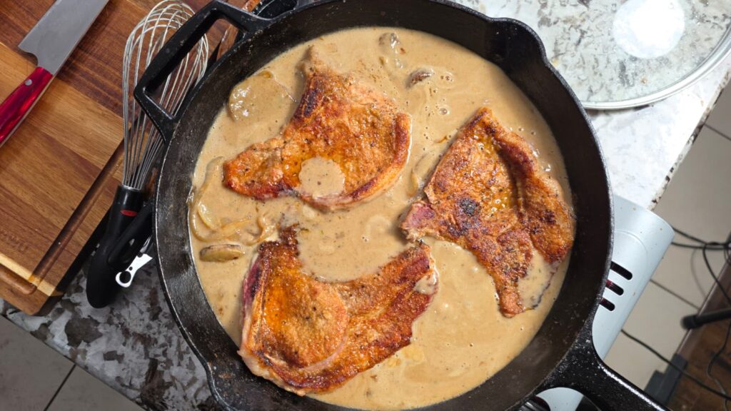 Three browned pork chops simmer in a creamy sauce with mushrooms in a black cast iron skillet on a stovetop, evoking the hearty comfort of classic smothered pork chops and potatoes, next to a wooden cutting board and a metal whisk.