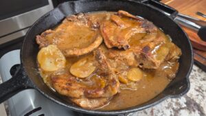 Smothered pork chops and potatoes simmer in rich brown gravy with sliced potatoes nestled in a black cast-iron skillet on a kitchen countertop.