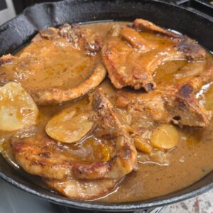 Smothered pork chops and potatoes simmer in rich brown gravy with sliced potatoes nestled in a black cast-iron skillet on a kitchen countertop.