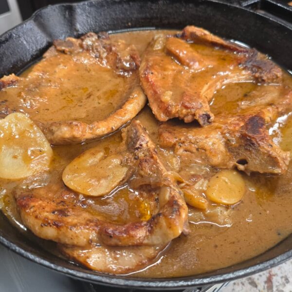 Smothered pork chops and potatoes simmer in rich brown gravy with sliced potatoes nestled in a black cast-iron skillet on a kitchen countertop.