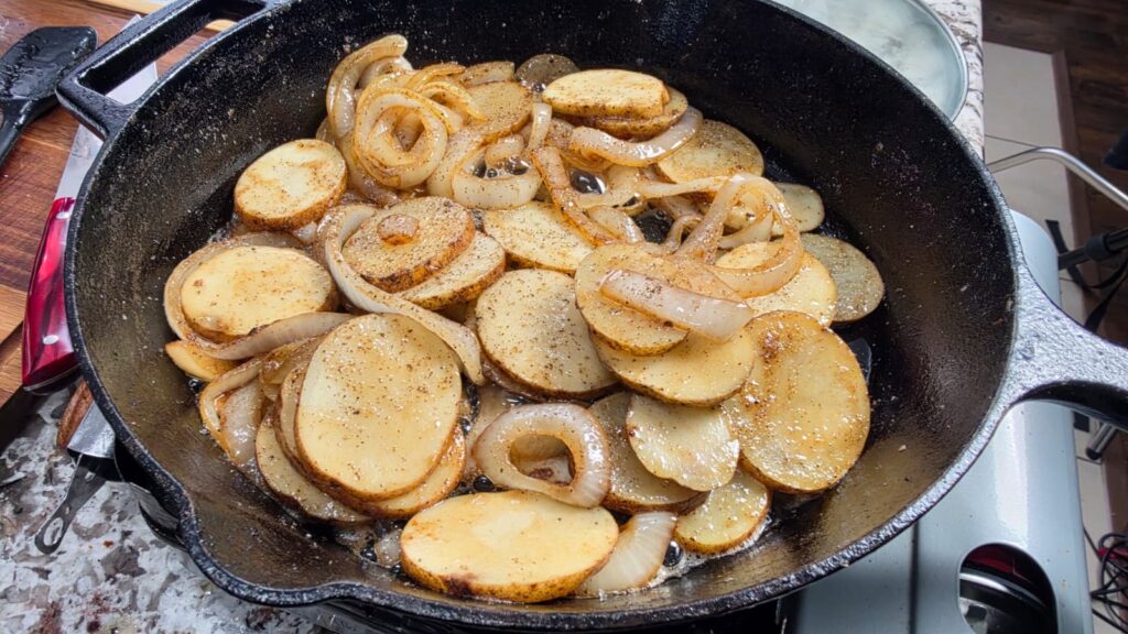 Sliced potatoes and onions are cooking in a black cast iron skillet, seasoned with pepper. The skillet is on a stove, and the vegetables are sizzling and starting to brown&mdash;perfect accompaniments for smothered pork chops and potatoes.