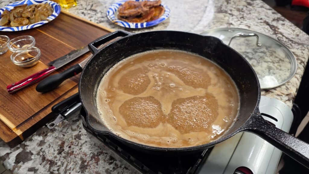 A cast iron skillet with bubbling brown gravy is cooking on a stovetop, filled with tender smothered pork chops and potatoes. Nearby are sliced meat on a paper plate, a cutting board with a knife, and small bowls of spices.
