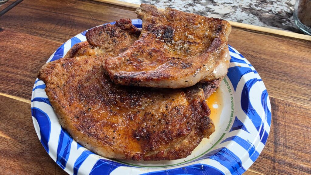 Two seasoned, cooked pork chops sit on a blue and white paper plate atop a wooden surface. The meat appears juicy with visible spices and grill marks, reminiscent of classic smothered pork chops and potatoes enjoyed at a comforting family dinner.