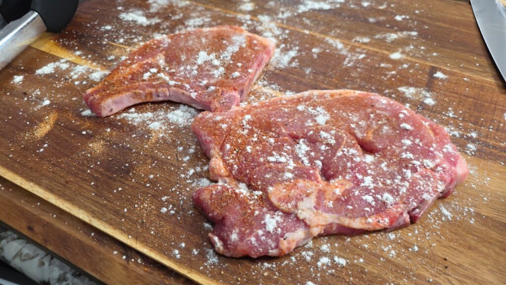 Two raw pork chops sprinkled with flour and seasoning rest on a wooden cutting board, ready to be transformed into savory smothered pork chops and potatoes. Some flour is scattered around the meat, while a knife and metal meat tenderizer are partially visible at the edges.