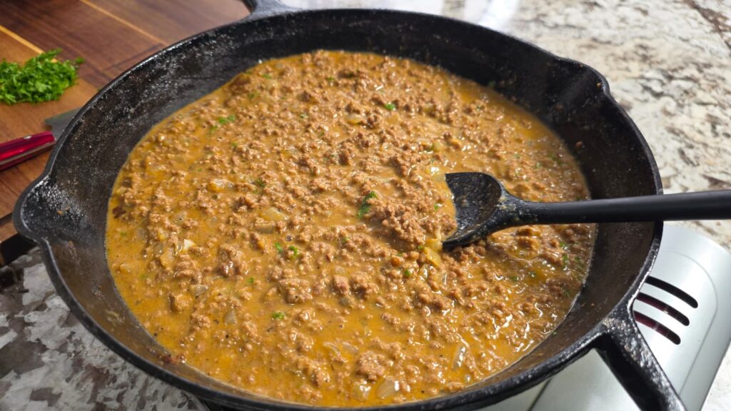 A cast iron skillet filled with a creamy smothered hamburger over rice ground meat mixture is being stirred with a black spoon on a kitchen counter.