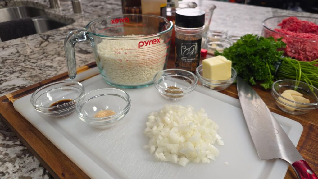 Chopped onions, spices, butter, and sauces are on a cutting board with a knife. Nearby are a measuring cup of rice, ground meat ready for smothered hamburger over rice, fresh parsley, and garlic on a kitchen counter.