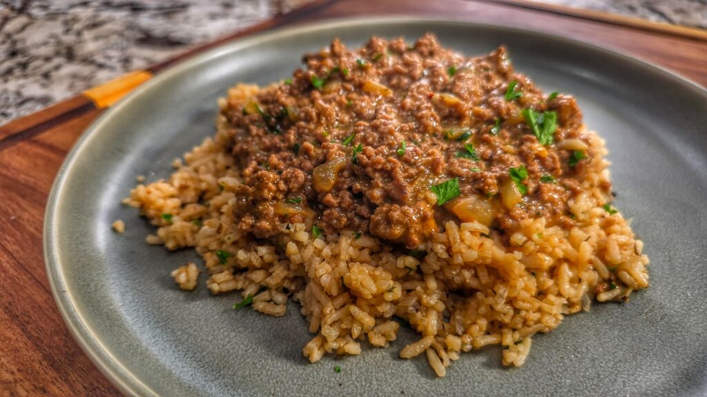 A plate of cooked rice topped with a savory, smothered hamburger and onion mixture, garnished with chopped herbs, served on a gray plate.