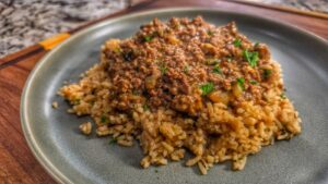 A plate of cooked rice topped with a savory, smothered hamburger and onion mixture, garnished with chopped herbs, served on a gray plate.