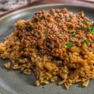 A plate of cooked rice topped with a savory, smothered hamburger and onion mixture, garnished with chopped herbs, served on a gray plate.