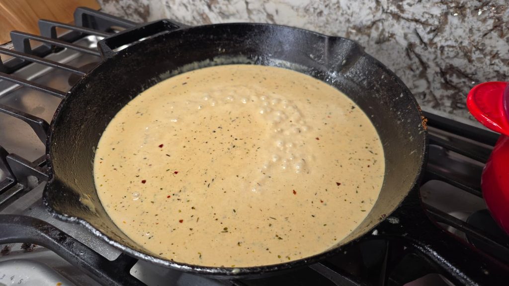 A cast iron skillet on a stovetop contains a creamy, bubbling garlic parmesan sauce with visible herbs and spices mixed in, perfect for smothering tender pork chops.