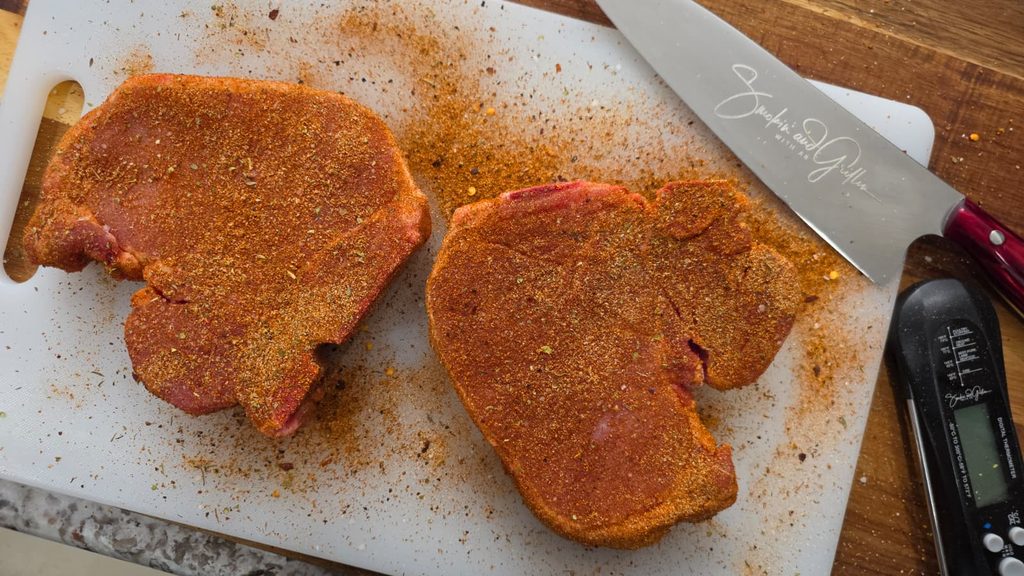 Two raw steaks coated with seasoning sit on a white cutting board, capturing the savory essence of garlic parmesan pork chops. A large knife and a digital meat thermometer are placed nearby, while spices are scattered around the board.