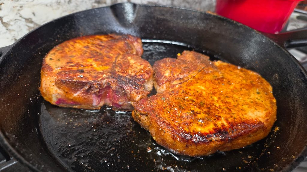 Two seasoned garlic parmesan pork chops are sizzling in a black cast iron skillet, showing a browned and crispy exterior, with a red pot blurred in the background.
