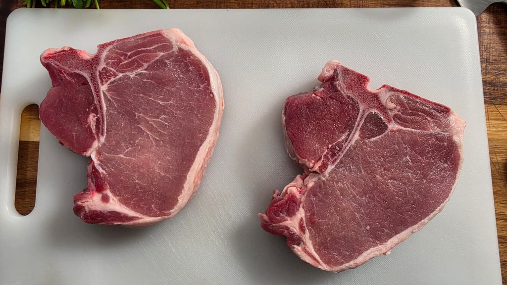Two raw garlic parmesan pork chops rest side by side on a white cutting board. The board sits on a wooden surface, and the uncooked meat is beautifully marbled, with a distinct T-bone showing in each chop.