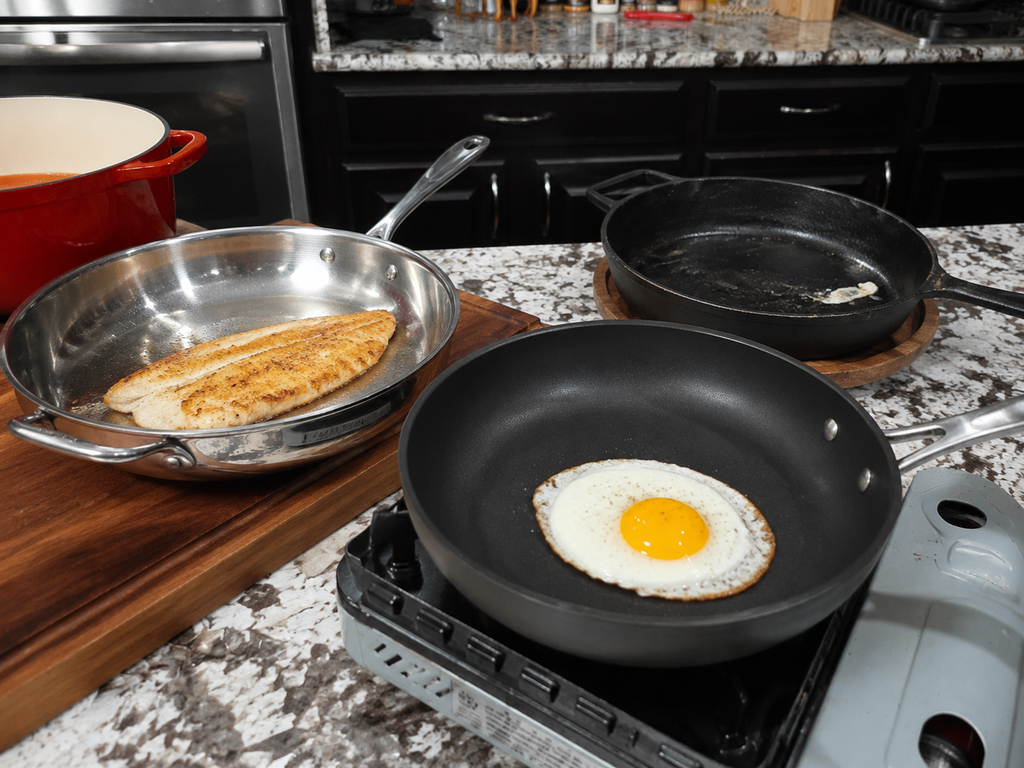 A fried egg cooks in a nonstick skillet on a stove, while a fillet of fish is searing in a stainless steel pan nearby. A cast iron pan sits empty on the counter. The kitchen is modern with dark cabinets.