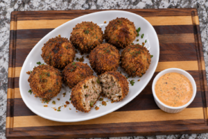 A white oval plate with eight breaded boudin balls, one cut open to show the flavorful interior, garnished with herbs. A small bowl of orange dipping sauce is on the side. Everything is arranged on a striped wooden board.