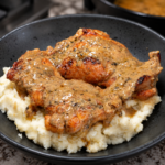 A black bowl filled with mashed potatoes topped with two pieces of Chicken Au Poivre, the roasted chicken covered in creamy, peppered gravy, sits on a granite countertop.