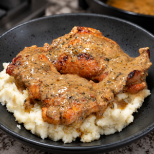 A black bowl filled with mashed potatoes topped with two pieces of Chicken Au Poivre, the roasted chicken covered in creamy, peppered gravy, sits on a granite countertop.