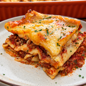 A slice of cheesy, baked Southern-style meat lovers lasagna with savory meat sauce is served on a white plate, garnished with chopped herbs. A casserole dish with more lasagna is in the background.
