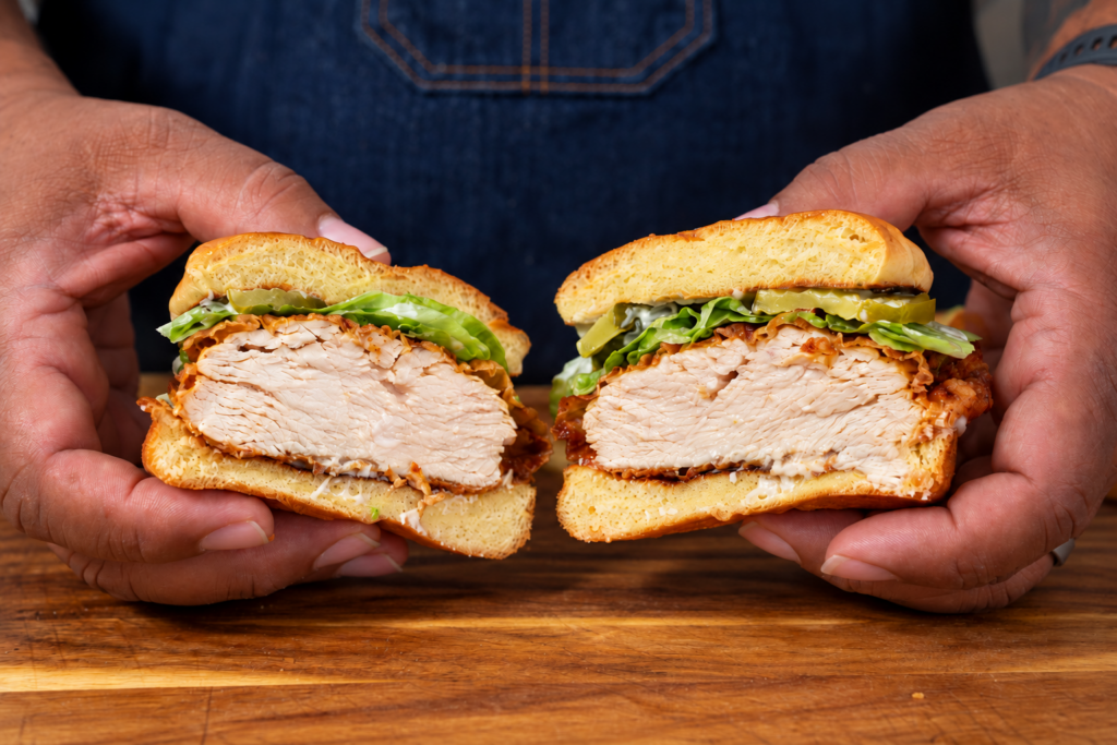 A person holds a sliced pickle brined fried chicken sandwich with lettuce and sauce on a wooden surface, revealing the juicy chicken inside.