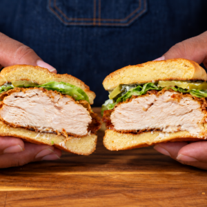 A person holds a sliced pickle brined fried chicken sandwich with lettuce and sauce on a wooden surface, revealing the juicy chicken inside.