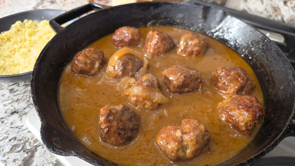 Meatballs simmer in brown gravy with onions in a black skillet. A plate of yellow rice sits nearby on a marble countertop.