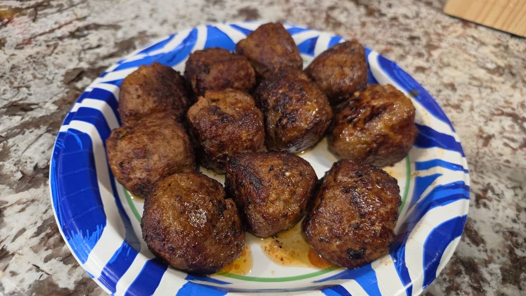 A paper plate with a blue and green pattern holds twelve browned meatballs on a granite countertop.