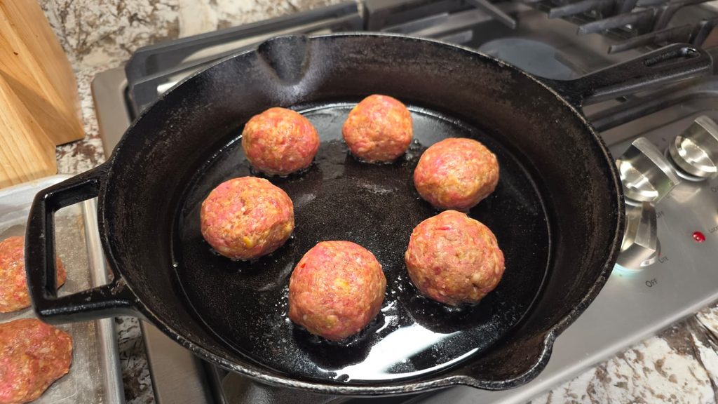 Six raw meatballs are arranged in a black cast iron skillet on a stovetop, with a few more meatballs visible on a tray nearby. The skillet has a bit of oil at the bottom.